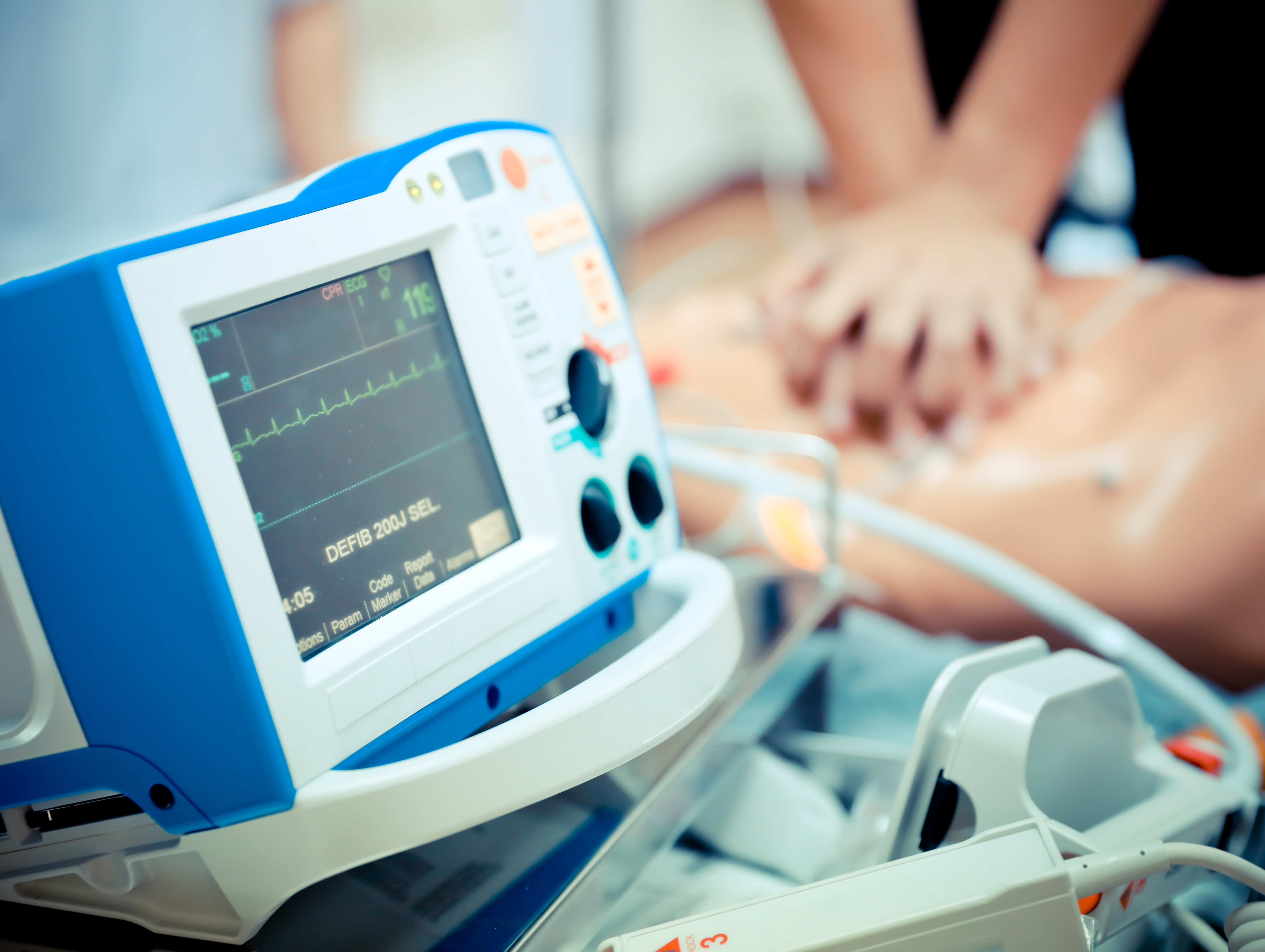 A defibrillator device, as part of a crash cart, sits alongside a nurse tending to a patient in cardiac arrest.